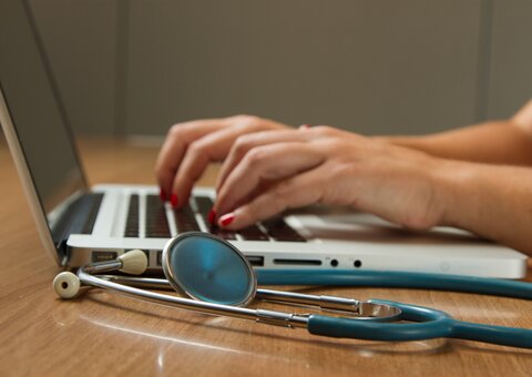 hands typing on a laptop, with a stethoscope on the table in the foreground