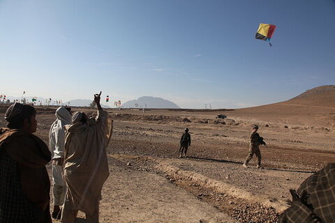 A group of people watching a person fly a kite against a blue sky, with two soldiers in the background