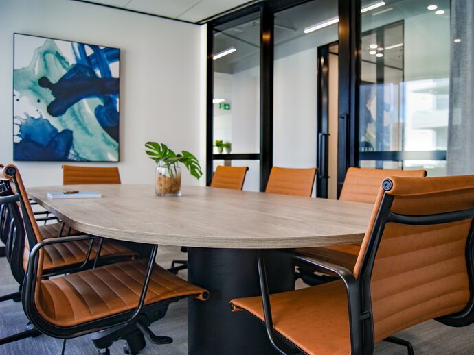 Photo of a meeting room, a blue painting is on the far wall and a large oval table is surrounded by black and orange chairs; a green plant in a pot sits on the table