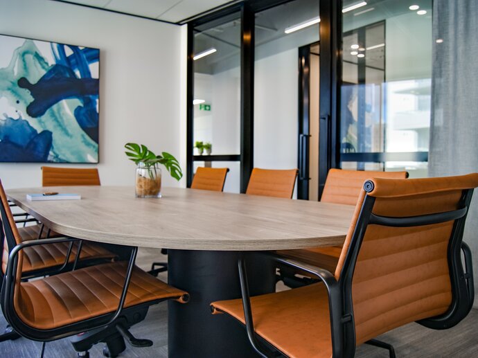 Photo of a meeting room, a blue painting is on the far wall and a large oval table is surrounded by black and orange chairs; a green plant in a pot sits on the table