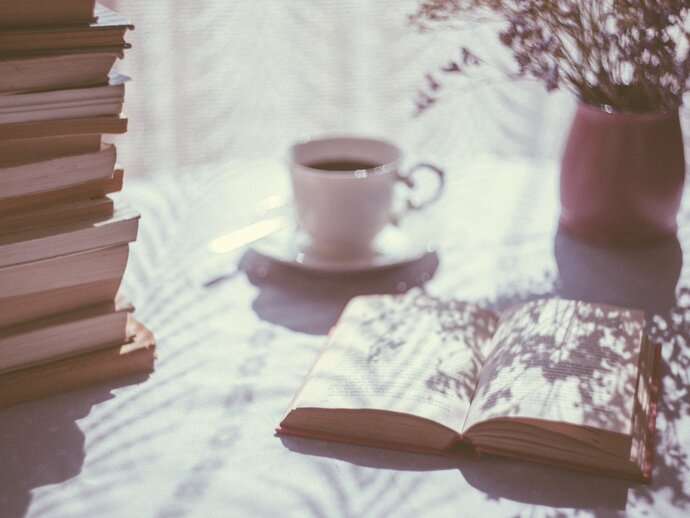 Photo of a stack of books, with one book laying open next to it, with a cup of tea and some flowers in the background.