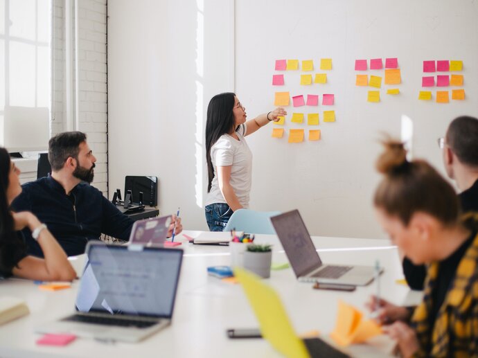 A group of people sitting around a meeting room table with laptops, watching a woman putting post-it notes on a wall