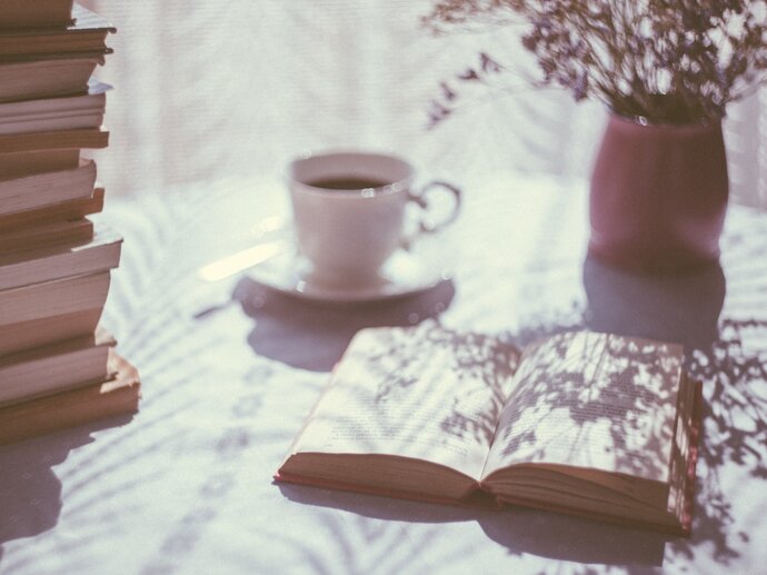 Photo of a stack of books, with one book laying open next to it, with a cup of tea and some flowers in the background.