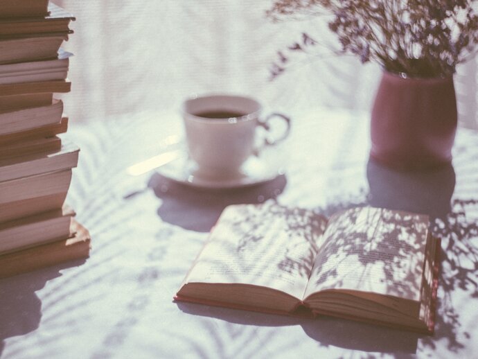 Photo of a stack of books, with one book laying open next to it, with a cup of tea and some flowers in the background.