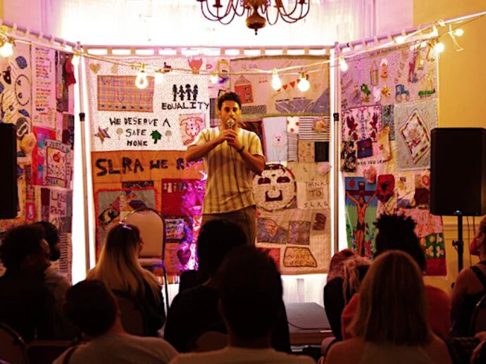 A man stands in front of a small group of people, speaking into a microphone. The wall behind him is covered in hand drawn art and writing