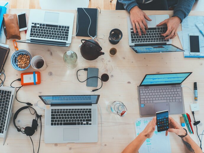 A group of laptops at a table, photographed from above. Several arms and hands indicate people working at the laptops 