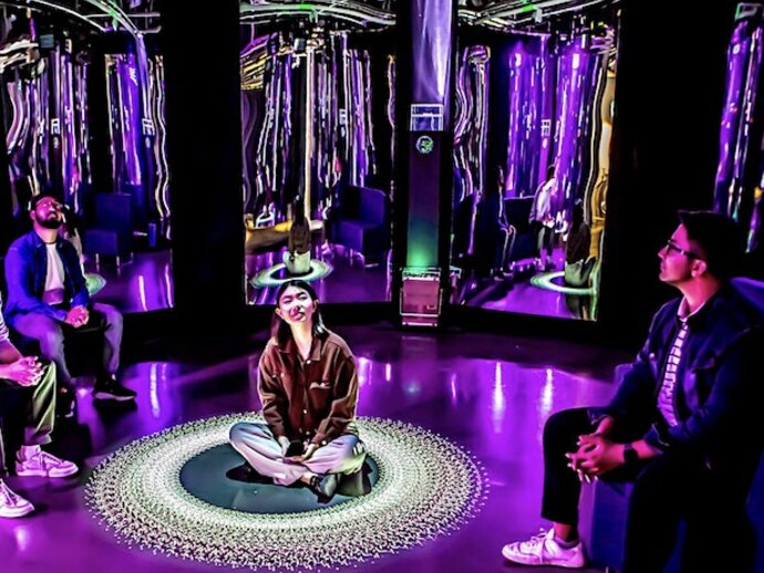 A woman sits in the middle of a room made of mirrors and purple lighting, there are other people sitting on chairs in the small room