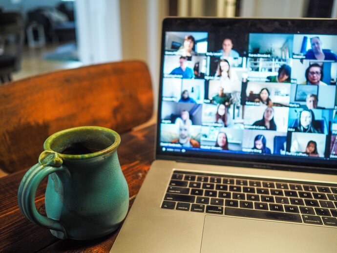 Online Co-production An open laptop displaying and online meeting screen with many attendees, a mug sits next to the laptop