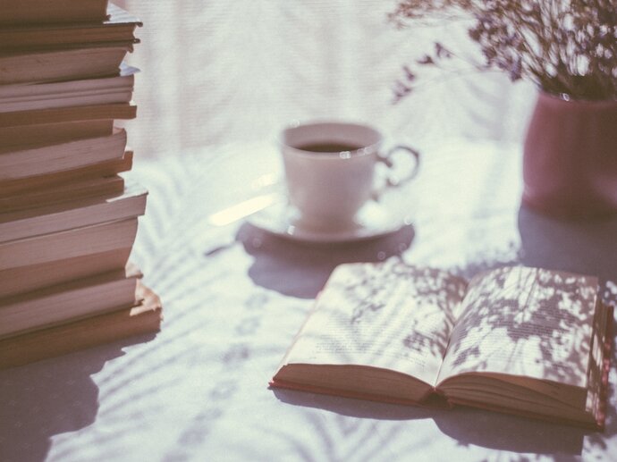 Photo of a stack of books, with one book laying open next to it, with a cup of tea and some flowers in the background.