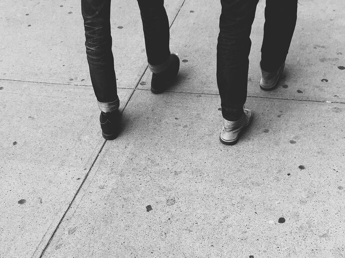 Two pairs of legs of people walking, the picture is cut off at the top so all you can see is legs and shoes on a concrete pavement.