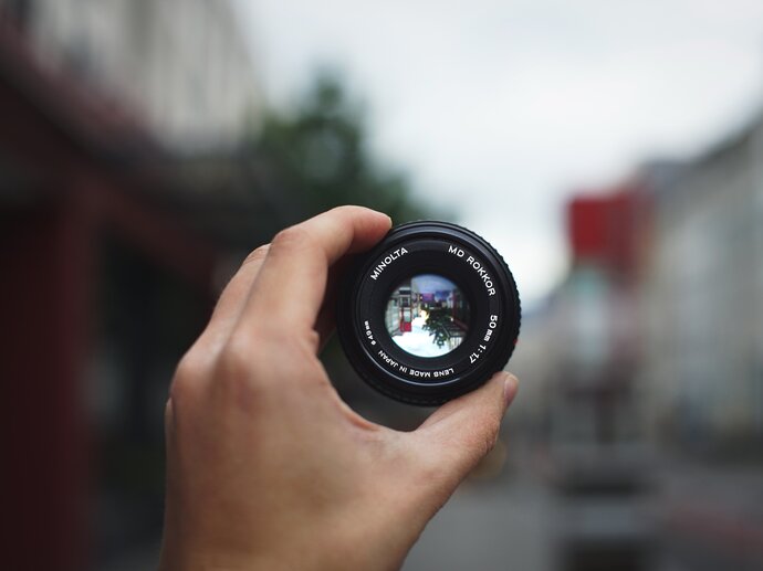 Picture of a hand holding up a camera lens over a blurred background, but through the lens everything is in focus