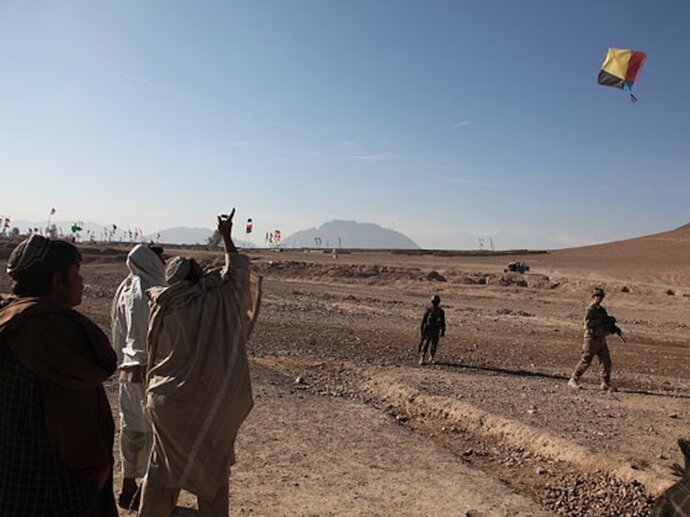 A group of people watching a person fly a kite against a blue sky, with two soldiers in the background