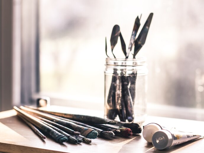 A jar of paintbrushes sits by a window, some paintbrushes and tubes of paint sit next to it. Reflecting the art-focused workshops discussed in the blog