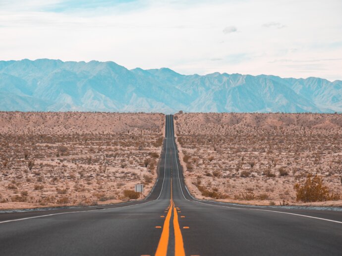 A long, straight road disappears into the distance through a flat dessert, mountains loom in the distance