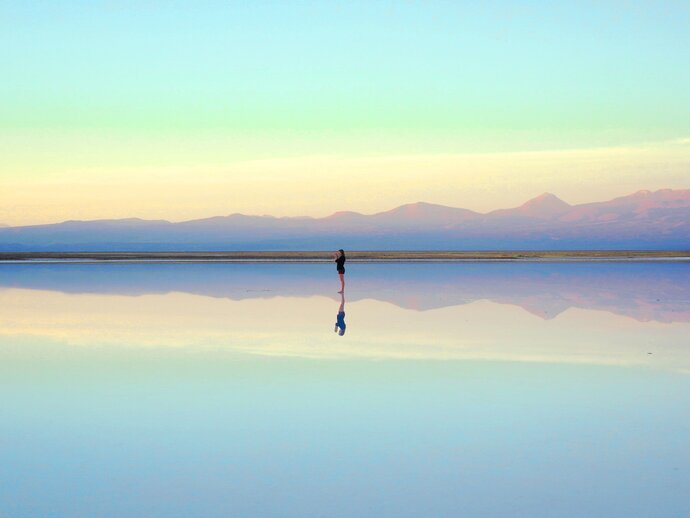A lone woman stands in shallow water in the centre of the image, with the mountains and pastel blue sky behind her perfectly reflected in the water