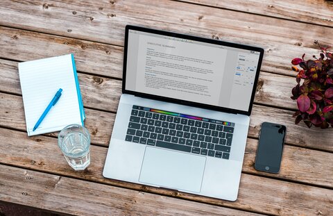 An open laptop sits on a wooden table, with a phone, glass of water, and notepad around it