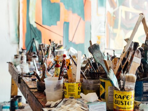 Photo of paintbrushes and art materials in a messy studio with a painting in the background