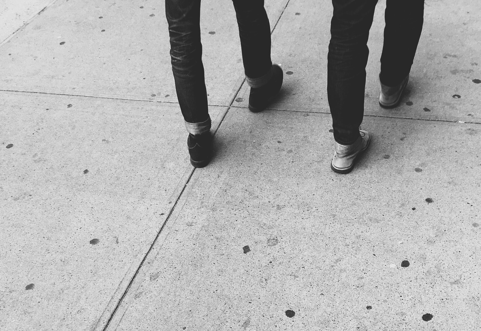 Two pairs of legs of people walking, the picture is cut off at the top so all you can see is legs and shoes on a concrete pavement.