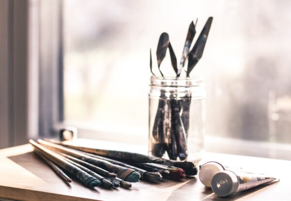 A jar of paintbrushes sits by a window, some paintbrushes and tubes of paint sit next to it. Reflecting the art-focused workshops discussed in the blog