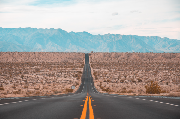 A long, straight road disappears into the distance through a flat dessert, mountains loom in the distance