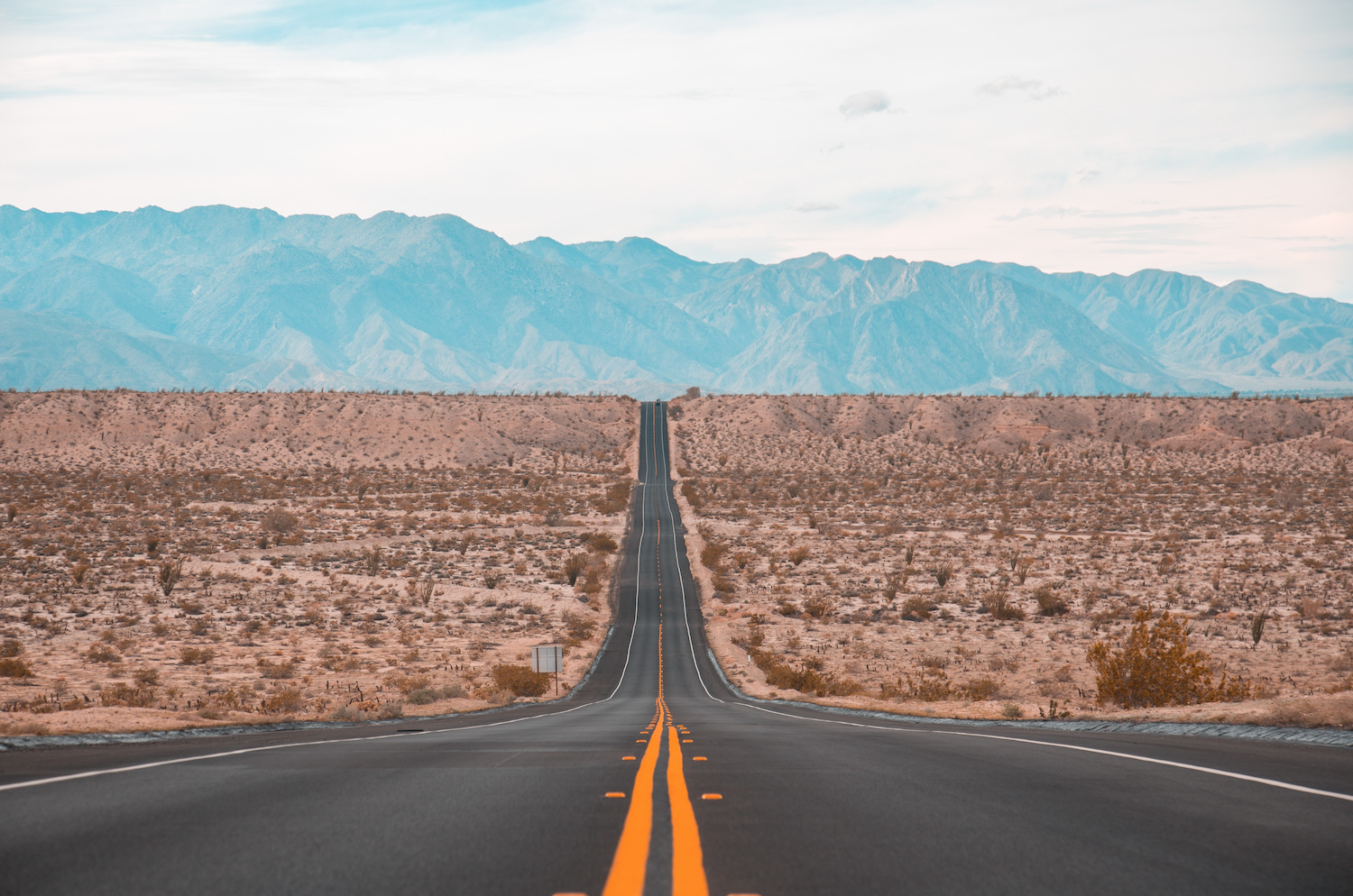 A long, straight road disappears into the distance through a flat dessert, mountains loom in the distance