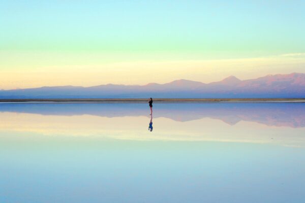 A lone woman stands in shallow water in the centre of the image, with the mountains and pastel blue sky behind her perfectly reflected in the water