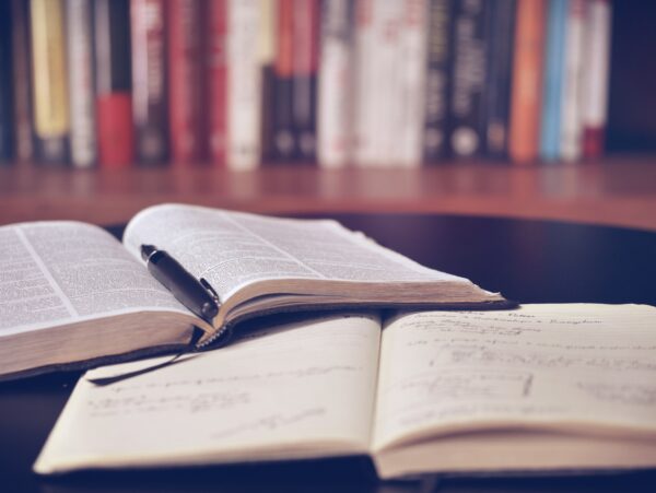 Photo of open books and notes with a pen, with library shelves in the background.