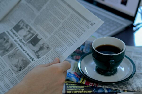 A person holding a newspaper with a cup of coffee on the table.